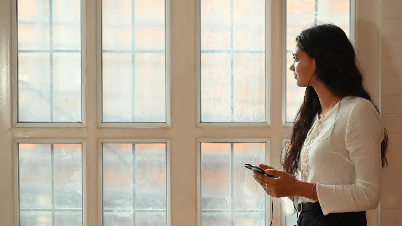 A woman standing by a window holding her phone