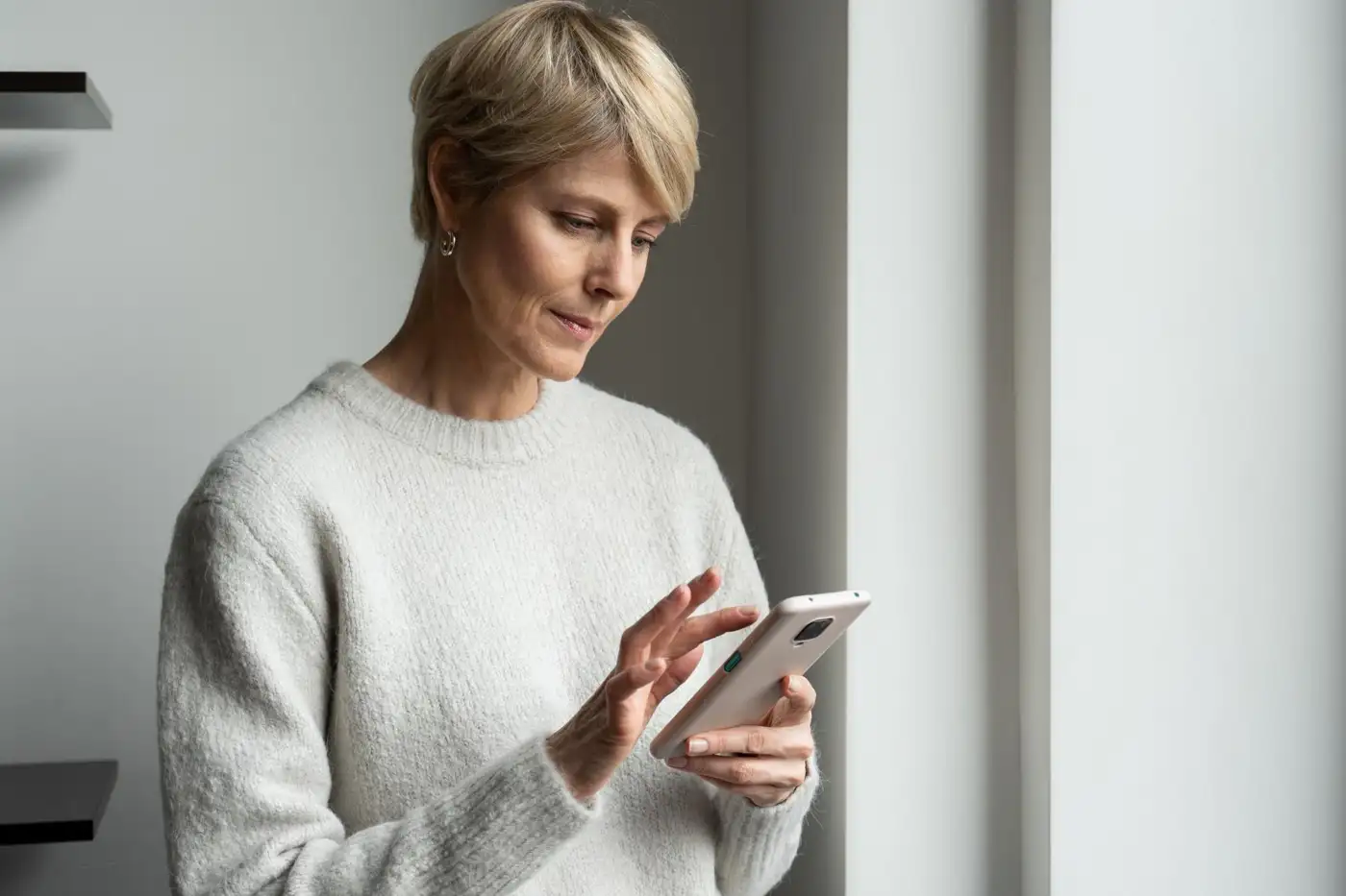 A woman standing by a window at home looking at her phone