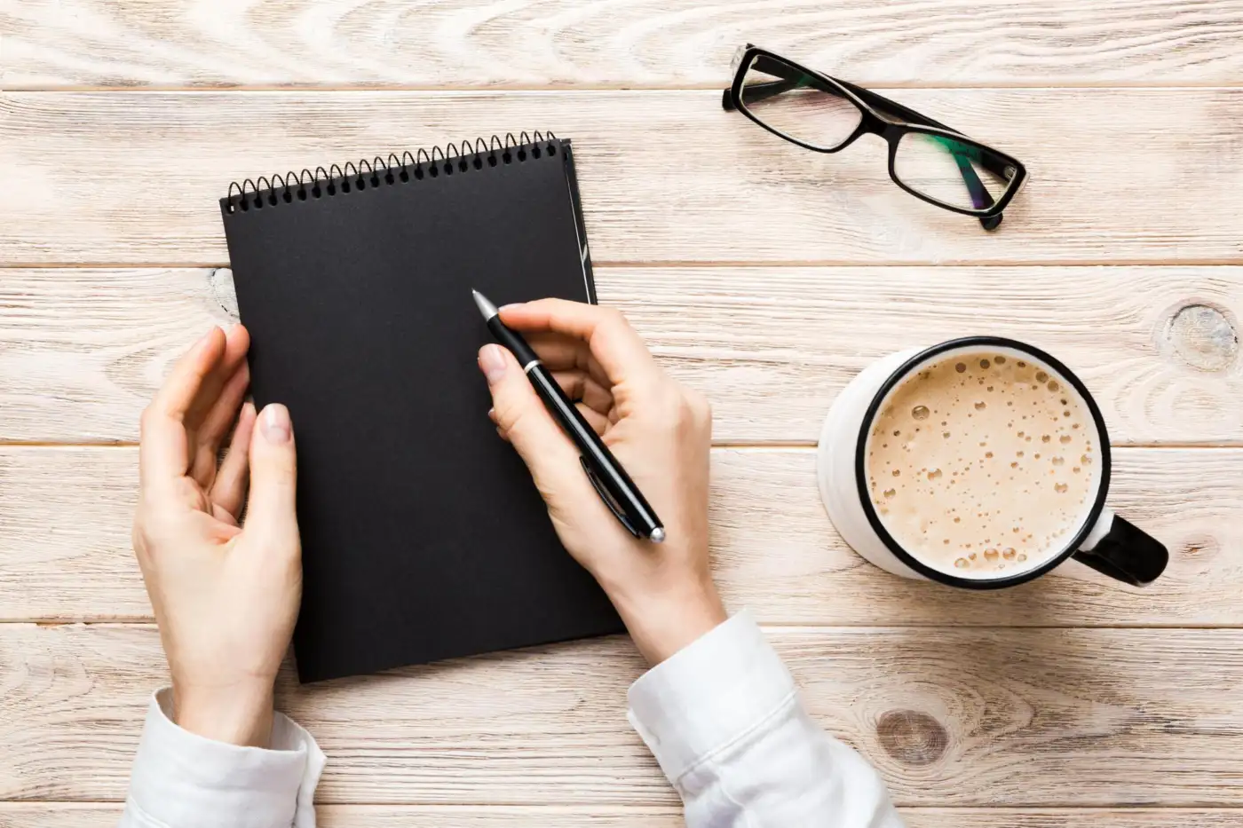 Hands writing in a notebook beside a mug of coffee