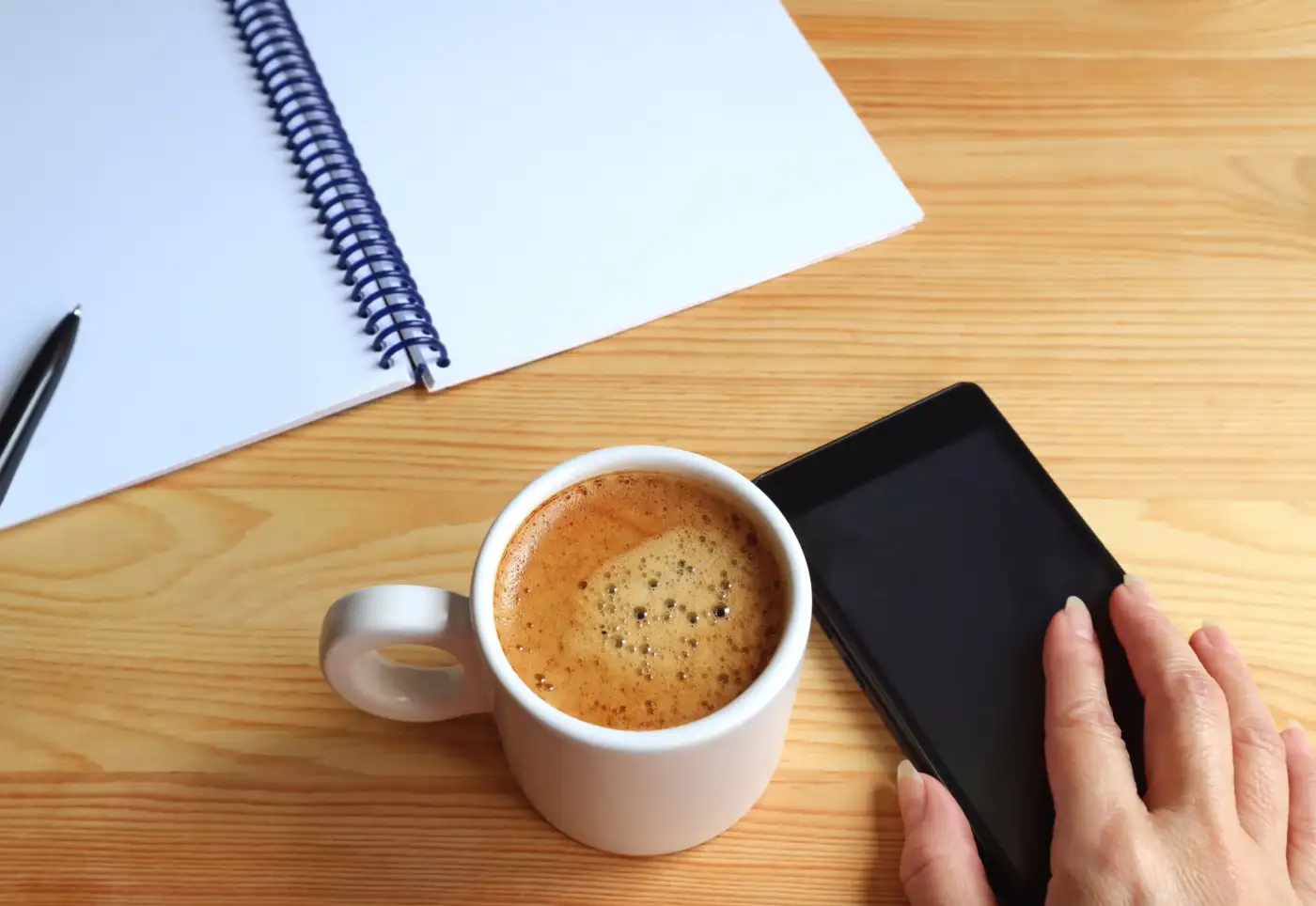 A notepad, a cup of coffee and a phone on a wooden table