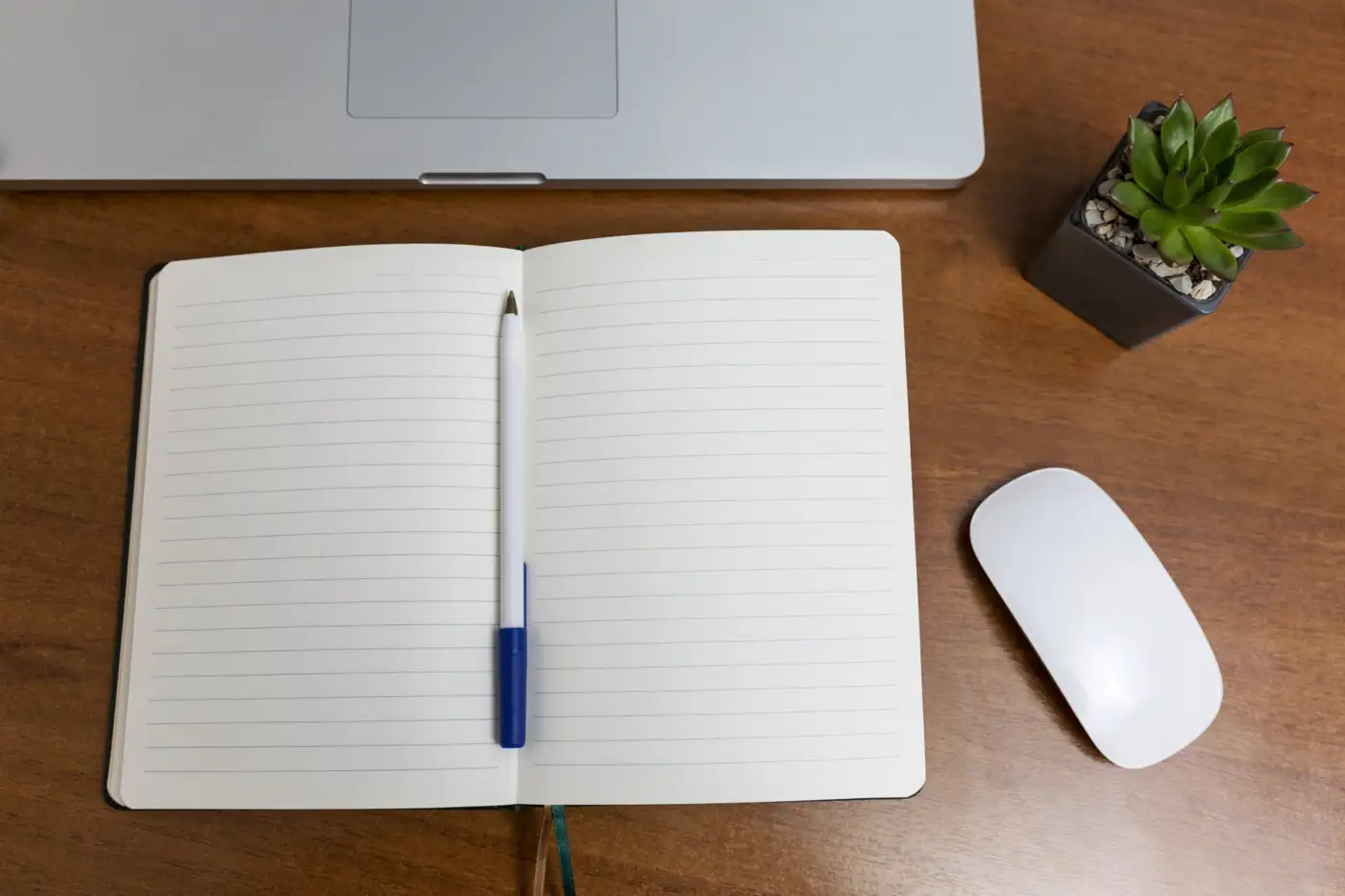 An open notebook and pen beside a laptop on a wooden desk