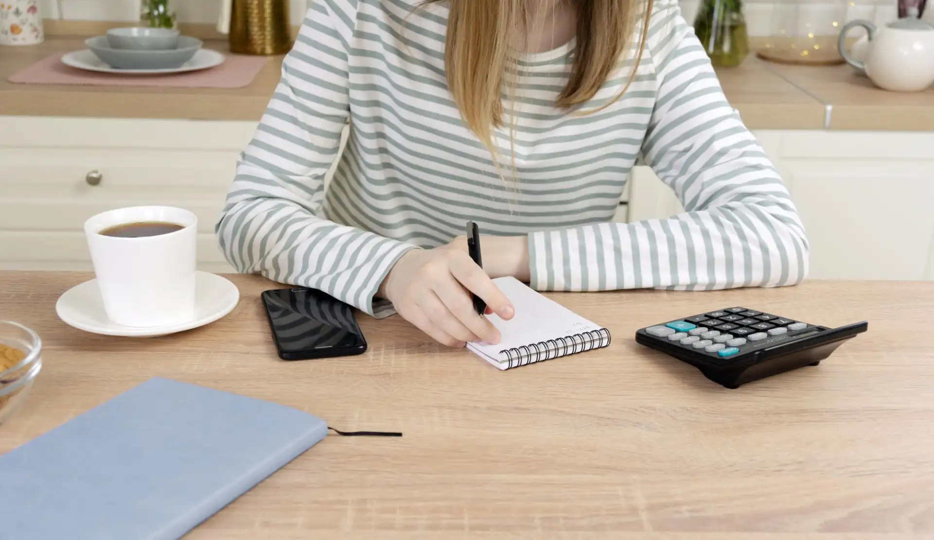 Hands writing in a notebook beside a calculator and a cup of coffee